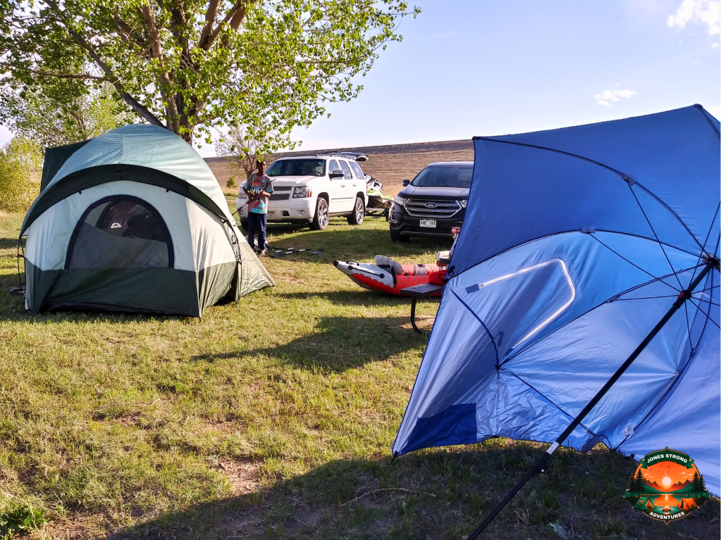 Camping setup with a green tent, blue canopy, cars, and an inflatable kayak on grassy land near a lake